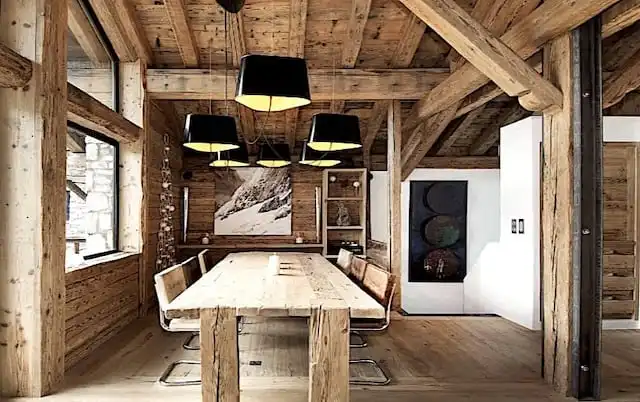 Une salle à manger rustique avec une table en bois, six chaises, des poutres en bois apparentes et cinq suspensions noires. La pièce dispose de grandes fenêtres, d'une photo de montagne sur le mur et met en valeur le lambris bois vieilli pour plus de charme.