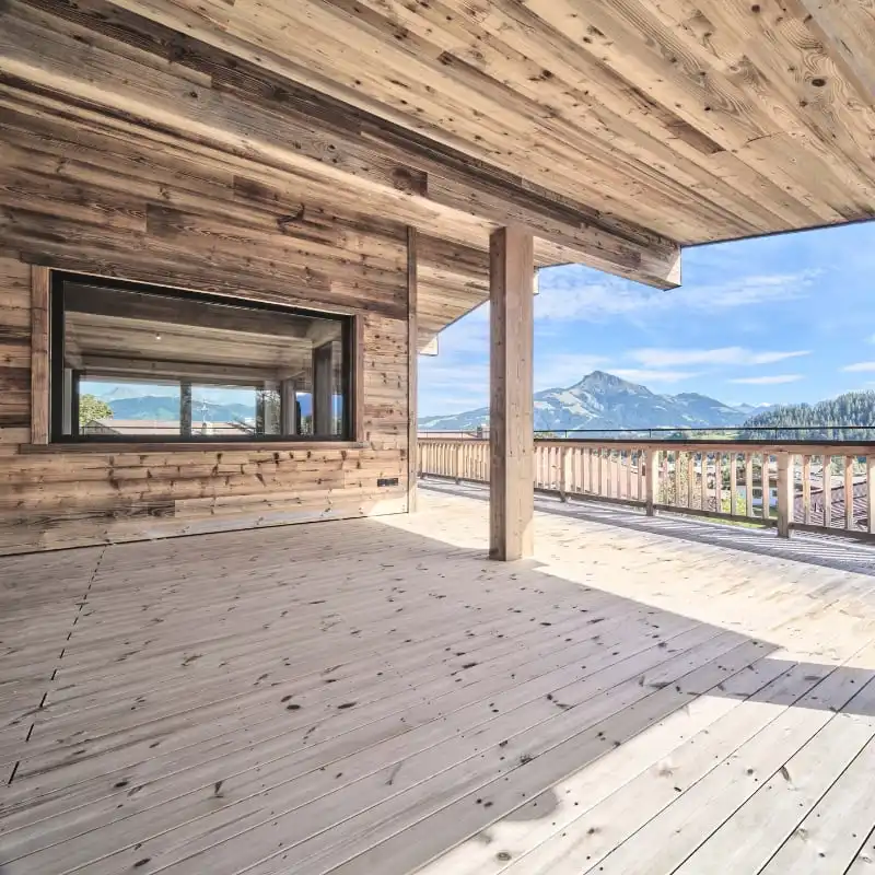  Terrasse et balcon en bois d'une maison comportant une grande fenêtre avec bardage vieux bois, donnant sur les montagnes et un paysage pittoresque sous un ciel bleu. 