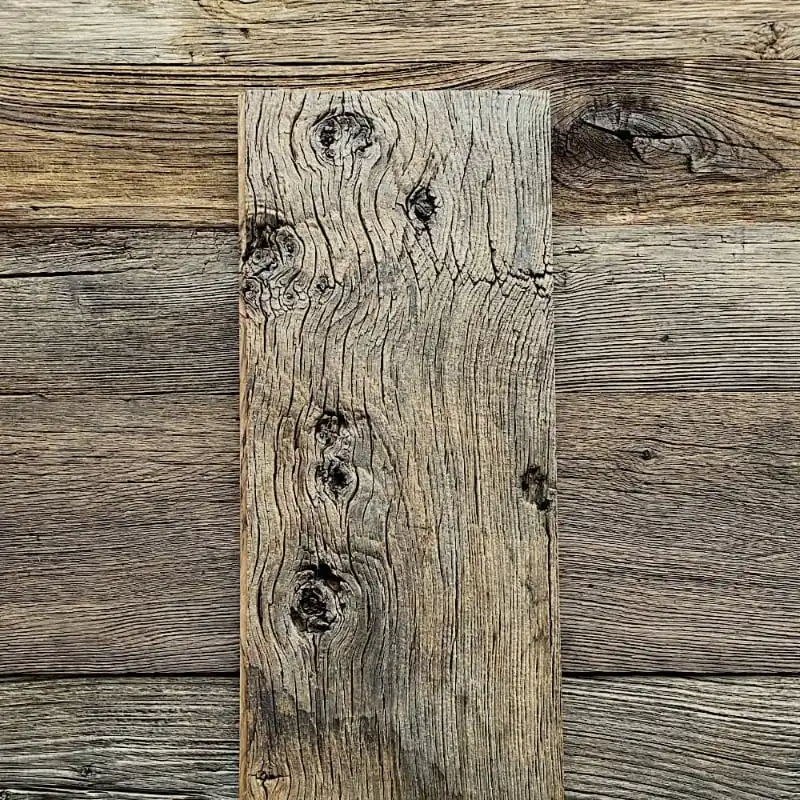  Un morceau de planche de bois gris patiné avec des nœuds et des veines visibles repose sur un fond de bardage châtaignier ancien tout aussi rugueux et aligné horizontalement. 