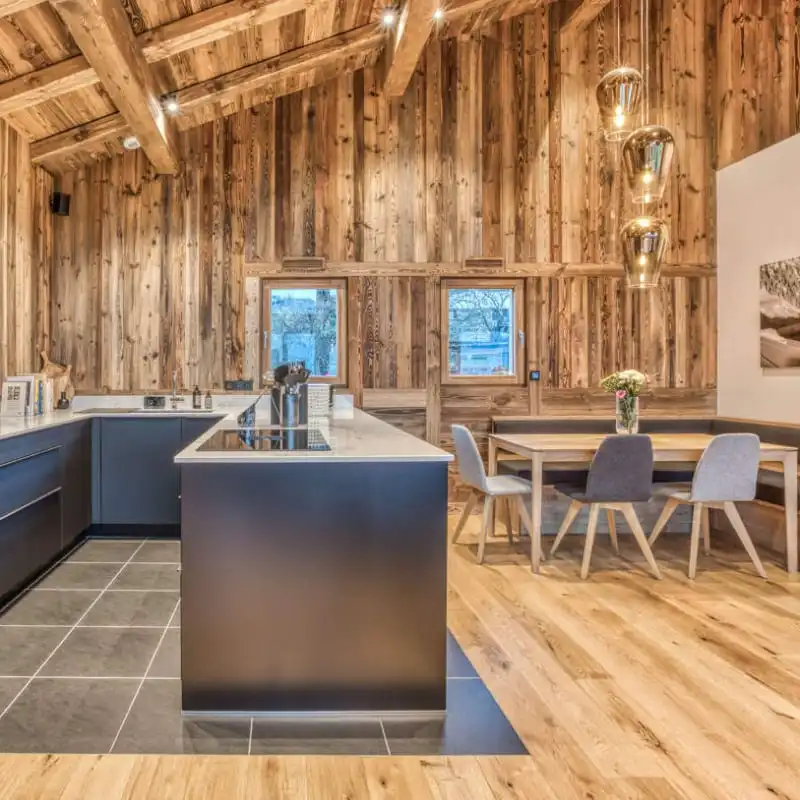  Cuisine moderne en bois avec de hauts plafonds, un îlot central avec des armoires foncées, un coin repas avec une table en bois et des chaises grises et des luminaires suspendus. L'accent de bardage bois ancien ajoute une touche de charme rustique à l'atmosphère chaleureuse et accueillante.  