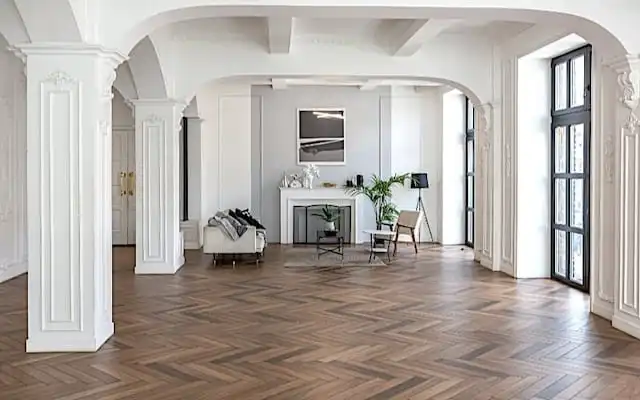 Un salon spacieux et élégant avec du parquet vieilli, des colonnes blanches, de grandes fenêtres et un mobilier minimaliste disposé autour d'une cheminée.