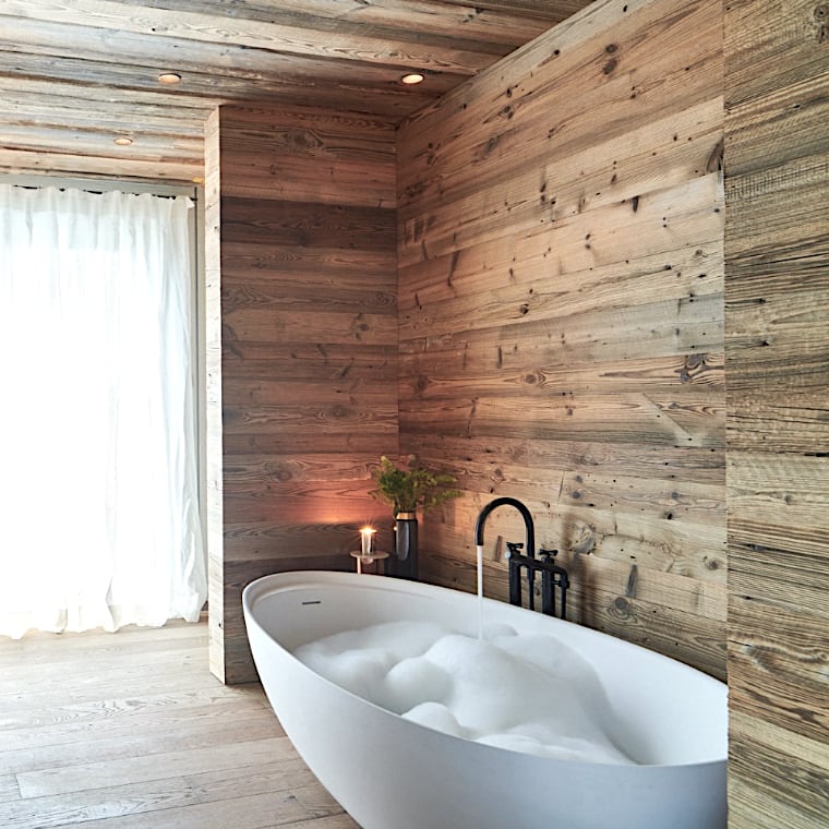 Une salle de bains moderne aux murs lambrissés, avec des touches de lambris bois ancien, met en valeur une baignoire autoportante blanche remplie de bulles. Une petite plante et une bougie sont placées à proximité, tandis qu'une fenêtre avec des rideaux transparents apporte de la lumière naturelle. 