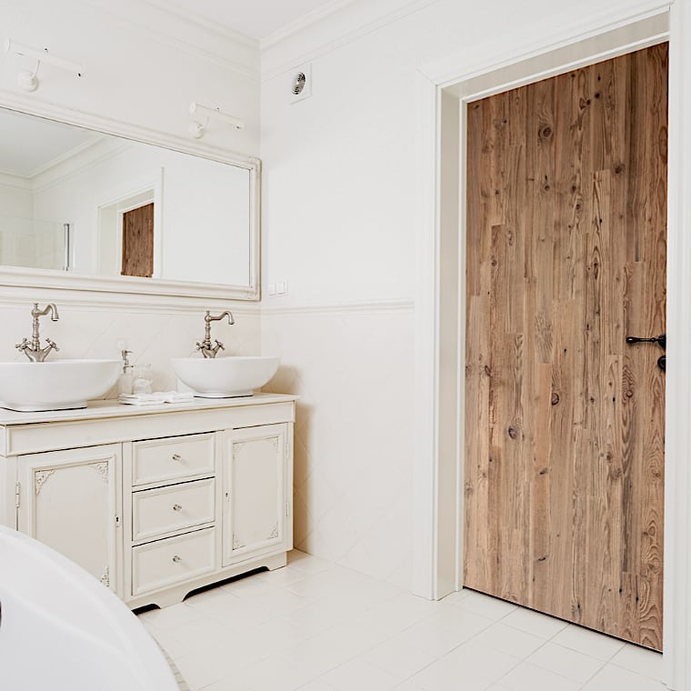  Une salle de bains lumineuse comprend une porte en bois rustique, des lavabos doubles avec des armoires ornées, de grands miroirs et un sol carrelé blanc. 
