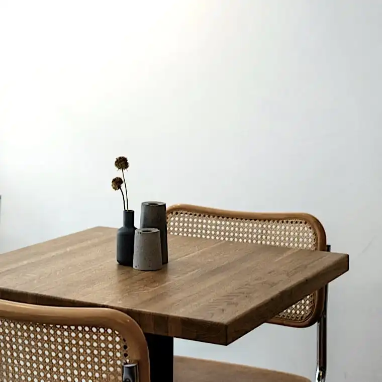  Un coin café minimaliste avec des tables et des chaises en bois dotées de dossiers et de sièges rustiques en osier. Une applique noire éclaire les murs en briques blanches et en plâtre. Un petit vase de fleurs séchées décore une table de bistrot. 