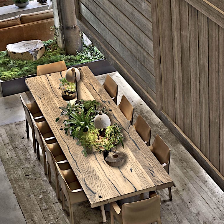  Une longue table à manger en bois, fond de chariot, avec dix chaises, est ornée de plantes en pot et d'objets décoratifs, située dans une pièce rustique et moderne avec des murs lambrissés et un jardin intérieur à proximité. 