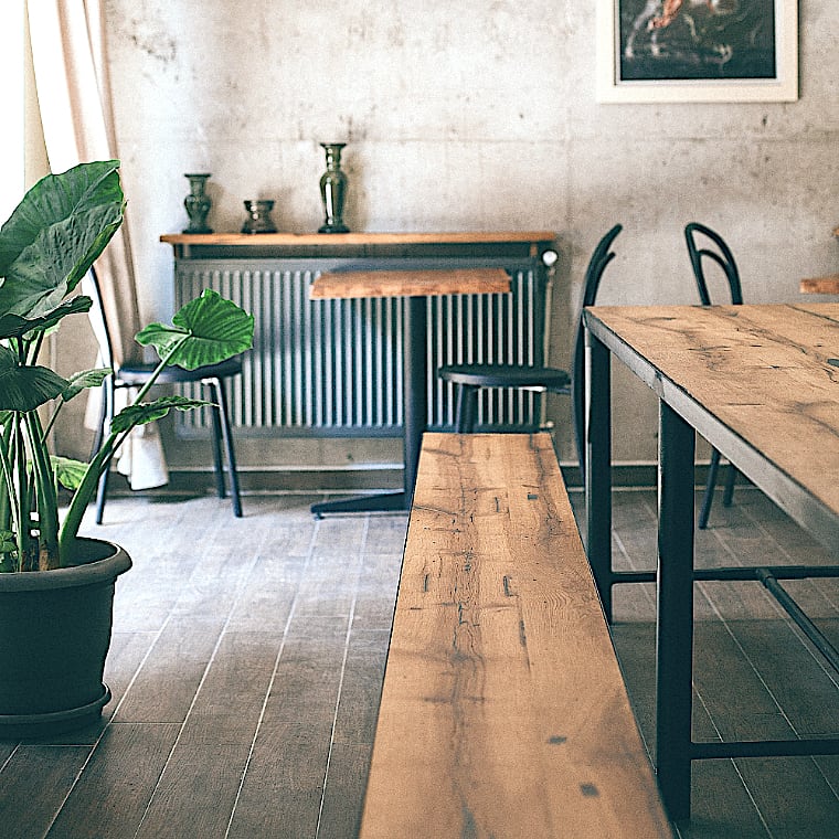  Une salle minimaliste avec des bancs, des tables et des chaises noires en bois. Une plante verte en pot repose sur le sol à côté d’une table et d’un banc bois recyclé. Une photo encadrée est accrochée au mur de béton. Parquet partout. 