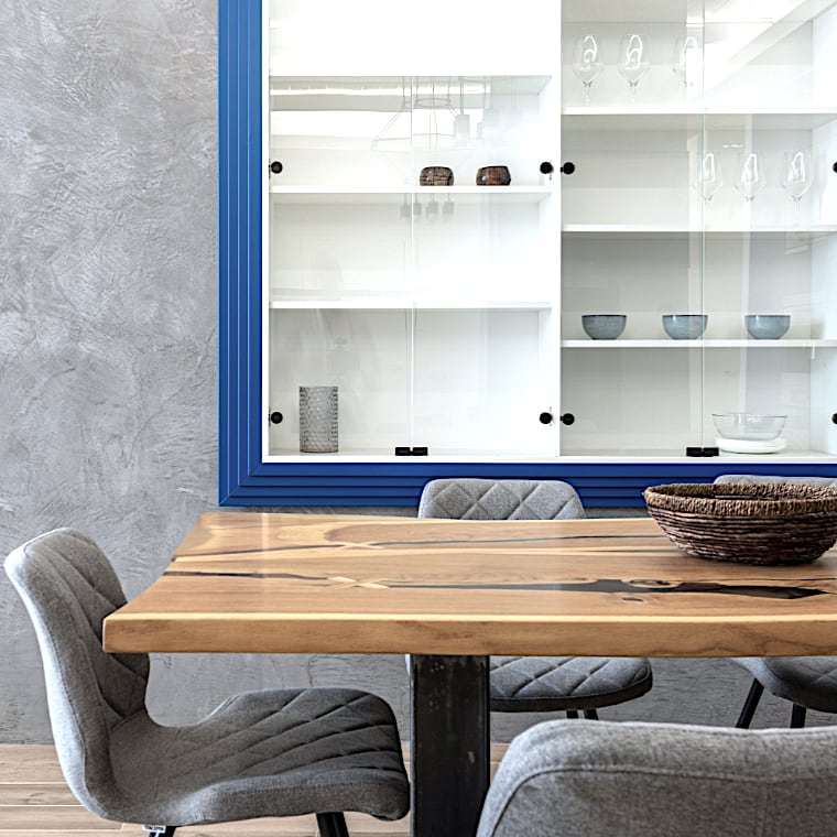  Une table à manger en bois recyclé avec des chaises rembourrées grises est placée devant une vitrine partiellement vide avec un cadre bleu, exposant quelques objets en verre et en céramique. 
