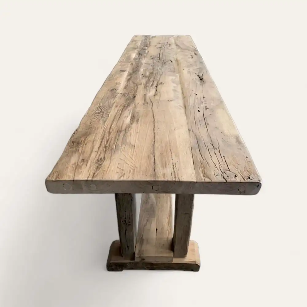  Une table rustique en bois avec un plateau épais et allongé et des pieds robustes en bois vieilli, rappelant une console en bois ancien, sur un fond neutre. 