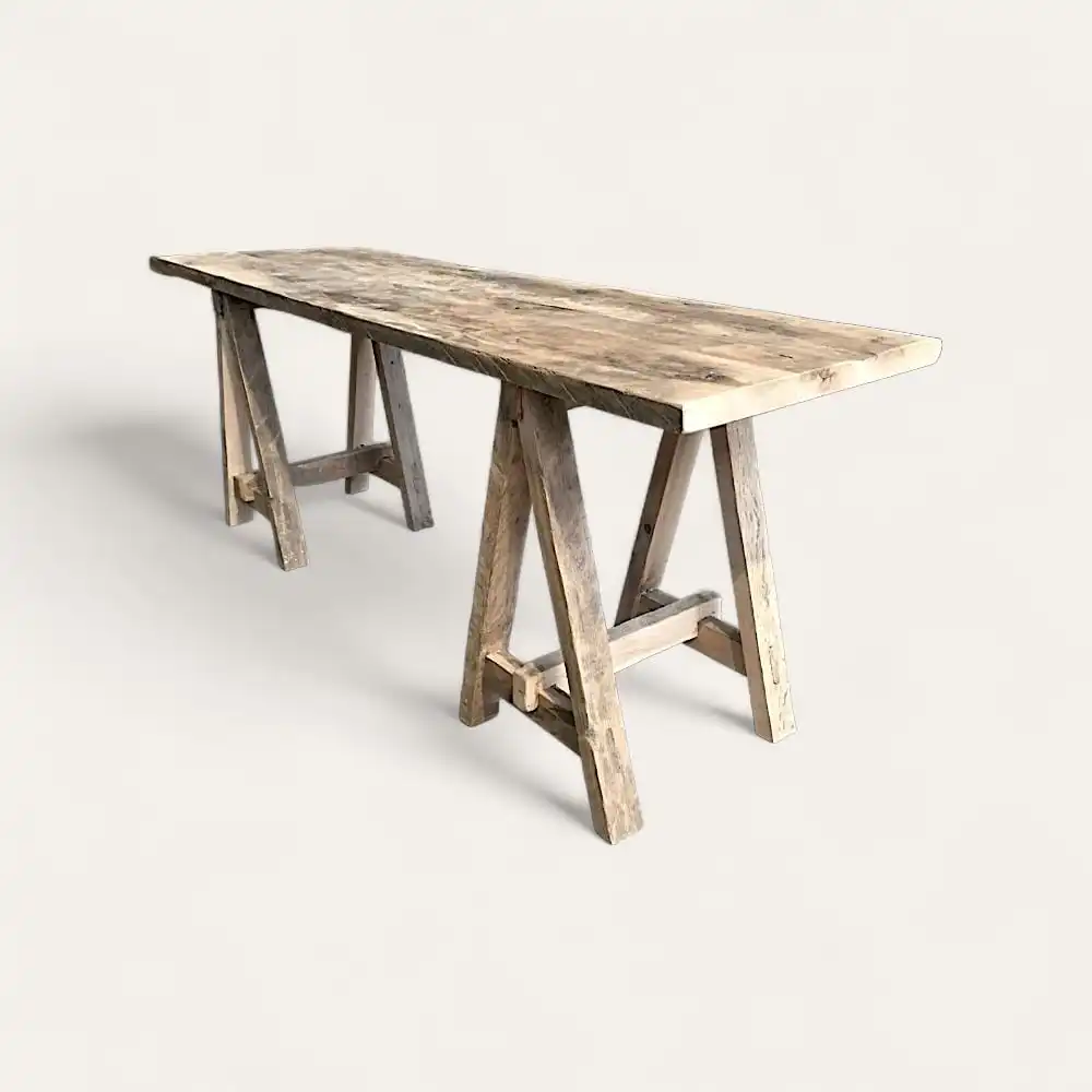  Une longue table en bois rustique avec un plateau rectangulaire et des pieds en tréteau, rappelant une console en bois ancien, est exposée sur un fond blanc uni. 
