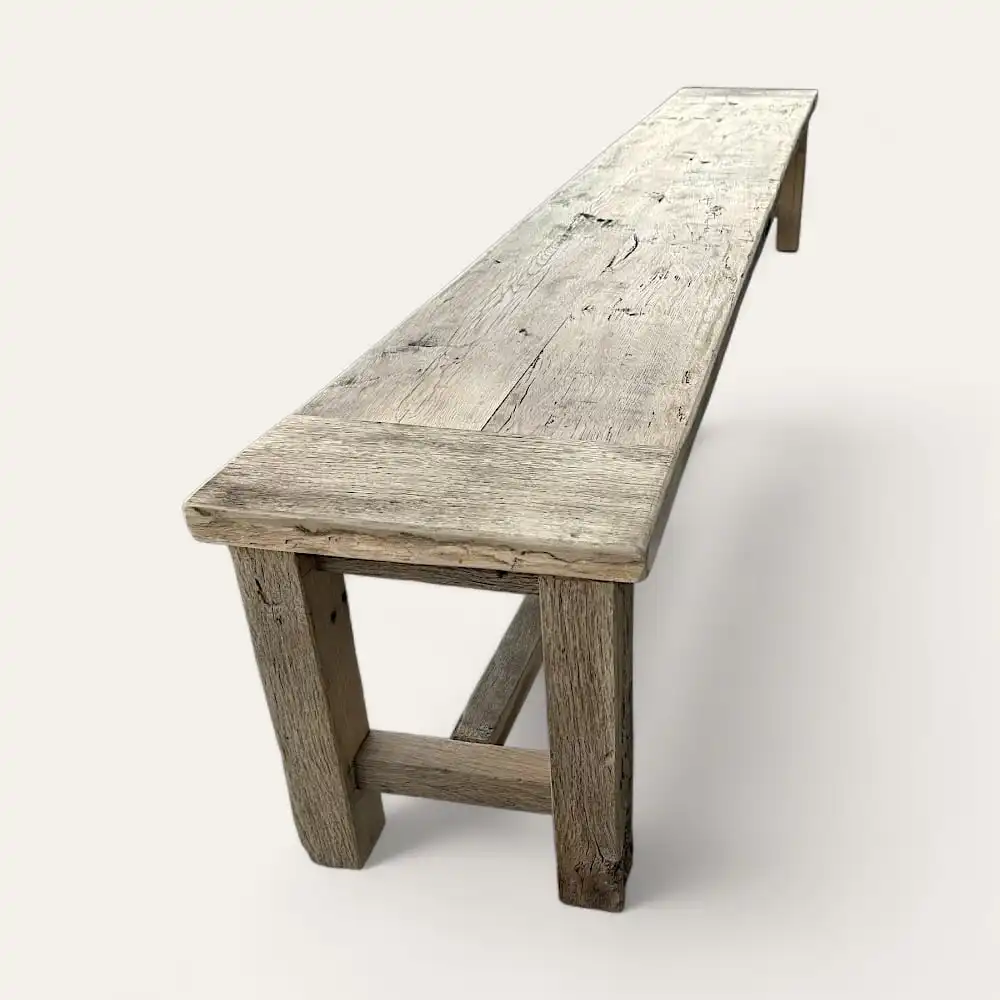  Un banc rustique en bois, ou banc rustique, avec un long siège en planches patinées et des pieds robustes. Le bois semble vieilli et texturé, lui donnant un aspect vintage. Le banc est placé sur un fond uni. 