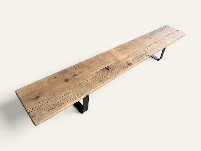 Un long banc bois ancien avec une finition rustique et deux pieds en métal noir sur fond uni.