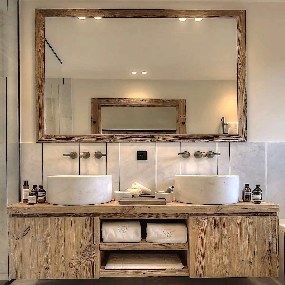  Salle de bain moderne comprenant un grand miroir avec cadre en bois, deux lavabos ovales sur une vanité à panneau 3 plis avec rangement et des murs en marbre. 