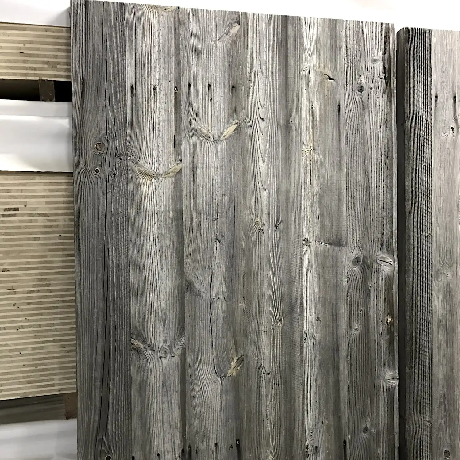  Panneaux de clôture en bois gris patiné, fabriqués à partir de vieux bois avec nœuds et grains visibles, posés contre un mur blanc. 