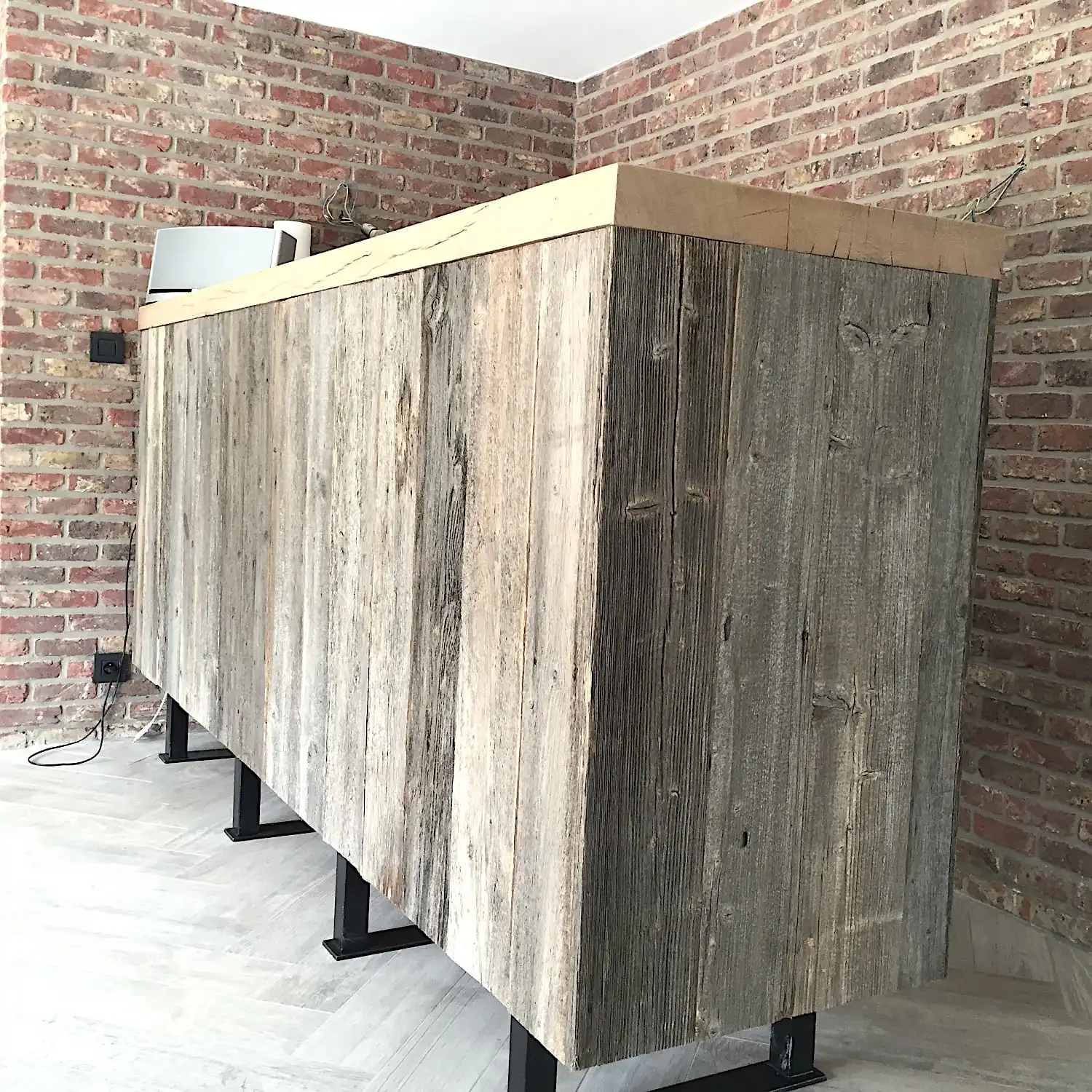  Un grand comptoir de bar en bois dans une pièce avec des murs en briques et une prise électrique visible, agrémenté de panneaux vieux bois. 