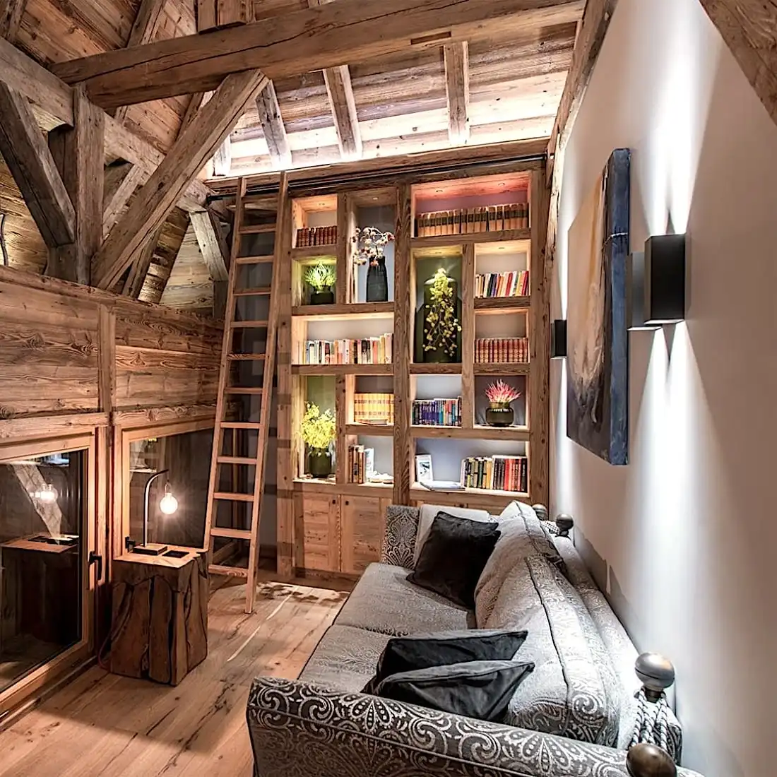  Chambre mansardée confortable avec poutres en bois, bibliothèque intégrée remplie de livres colorés, échelle, canapé confortable et éclairage tamisé grâce à des panneaux vieux bois. 