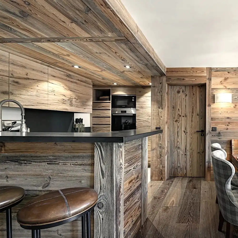  Intérieur d'une cuisine rustique avec des murs en bois et des armoires en panneaux de bois vieux, comprenant des appareils électroménagers modernes, un bar de petit-déjeuner avec des tabourets et une chaise rembourrée à carreaux. 