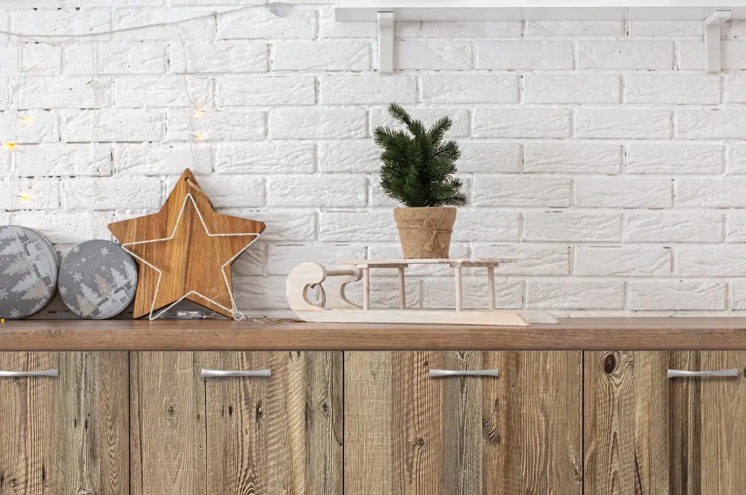 Une étagère en panneau 3 plis vieux bois ornée de décorations de Noël dont une étoile, un arbre miniature, un traîneau et des boules à motifs, contre un mur de briques blanches.