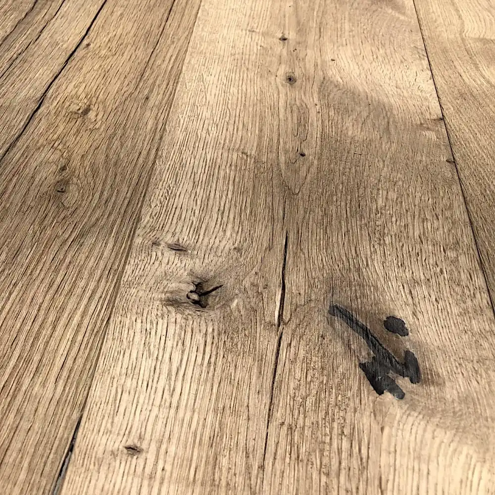  Gros plan d'une surface en bois avec des motifs de grain visibles, des nœuds et quelques marques sombres. La texture du plancher ancien est rugueuse et naturelle. 
