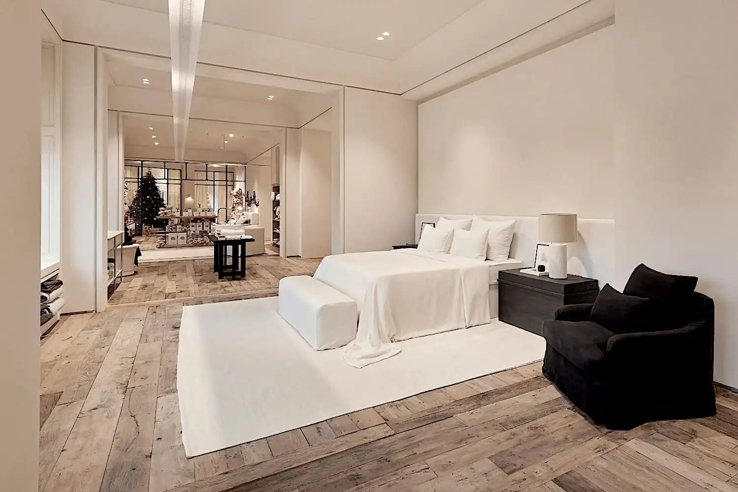 Une chambre spacieuse avec un grand lit blanc, des tables de chevet noires et un fauteuil au sol châtaignier. Un tapis se trouve sous le lit et un espace de vie ouvert est visible en arrière-plan.