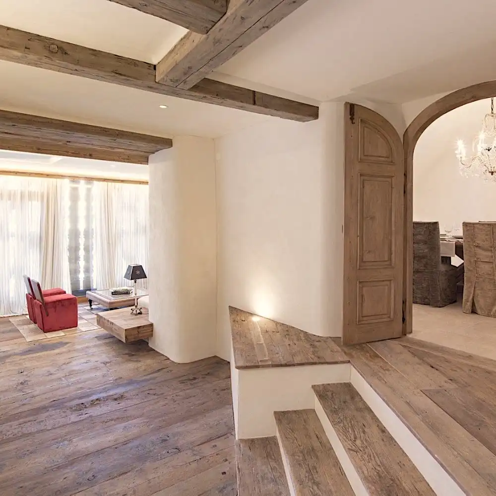  Un espace de vie lumineux avec des poutres en bois et du parquet ancien rustique comprend un coin salon avec une chaise rouge, une table en bois, des escaliers et une porte cintrée menant à un coin repas avec un lustre. 