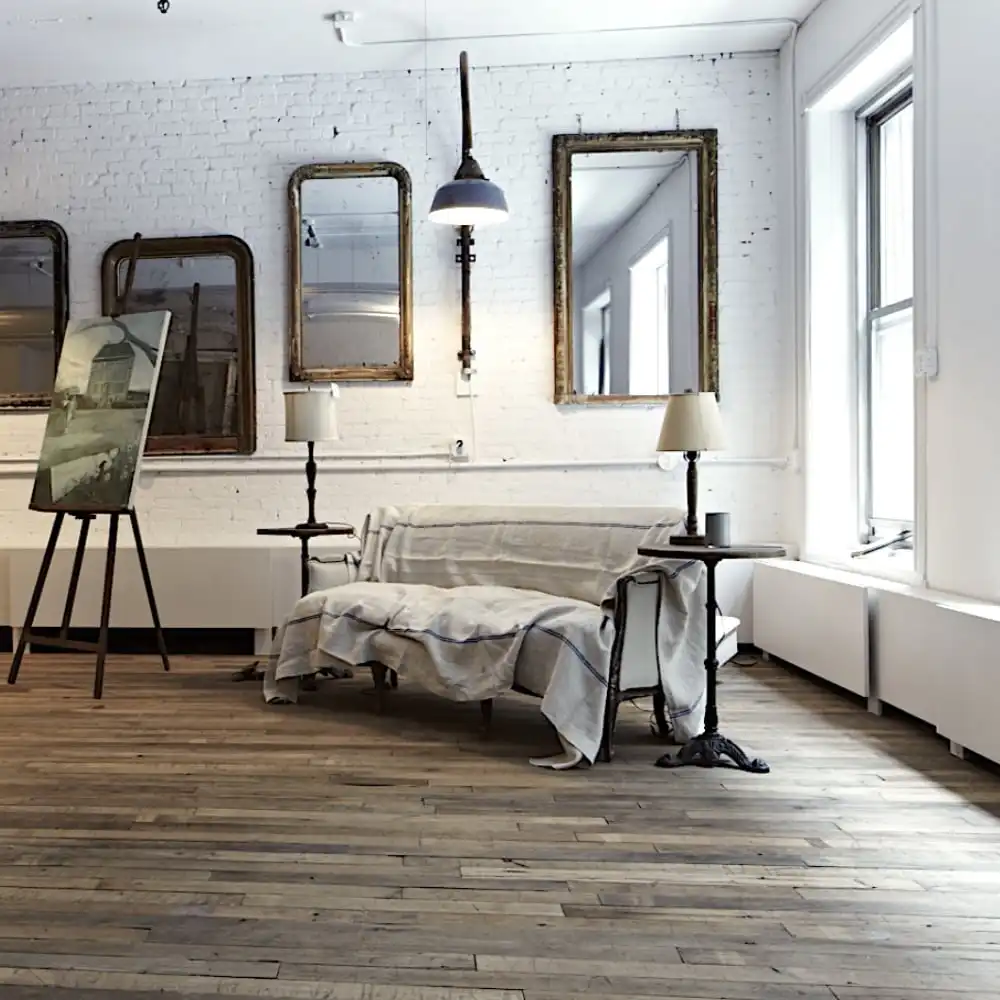  Une pièce minimaliste avec parquet ancien comprend un canapé couvert, deux tables d'appoint avec lampes, des miroirs encadrés sur un mur de briques et un tableau sur un chevalet près de la fenêtre. 