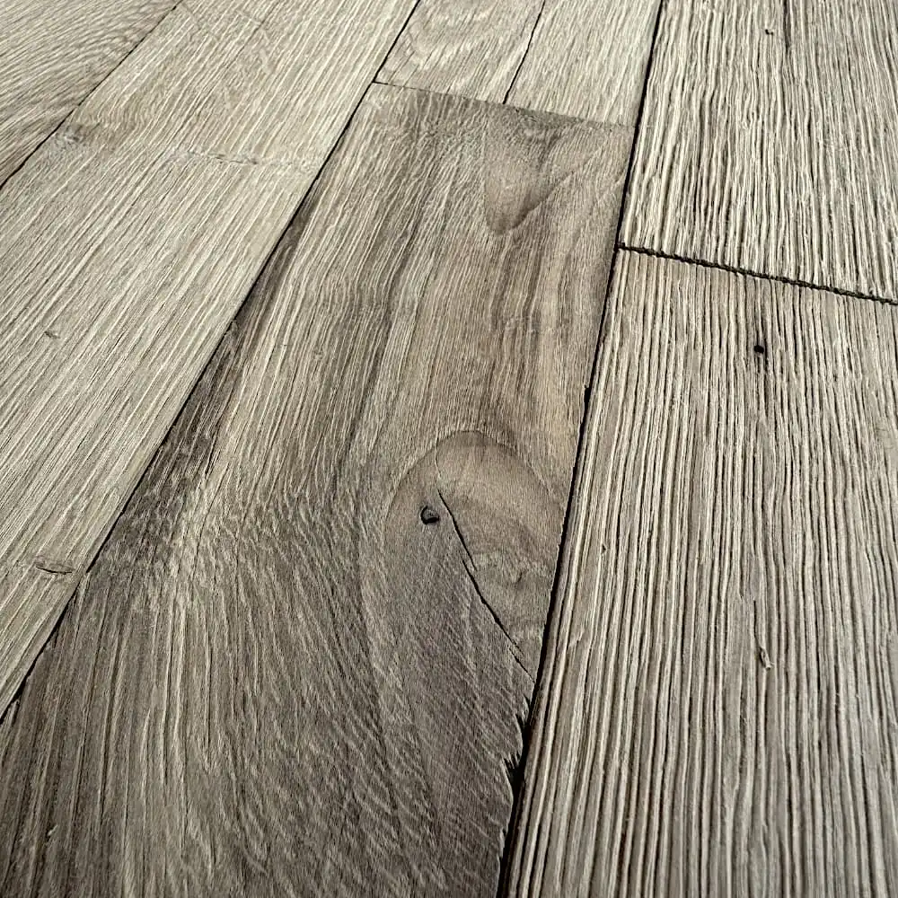  Gros plan de planches de bois texturées disposées selon un motif de revêtement de sol, présentant un mélange de motifs de grain et de nuances de brun et de gris, rappelant le parquet ancien classique. 