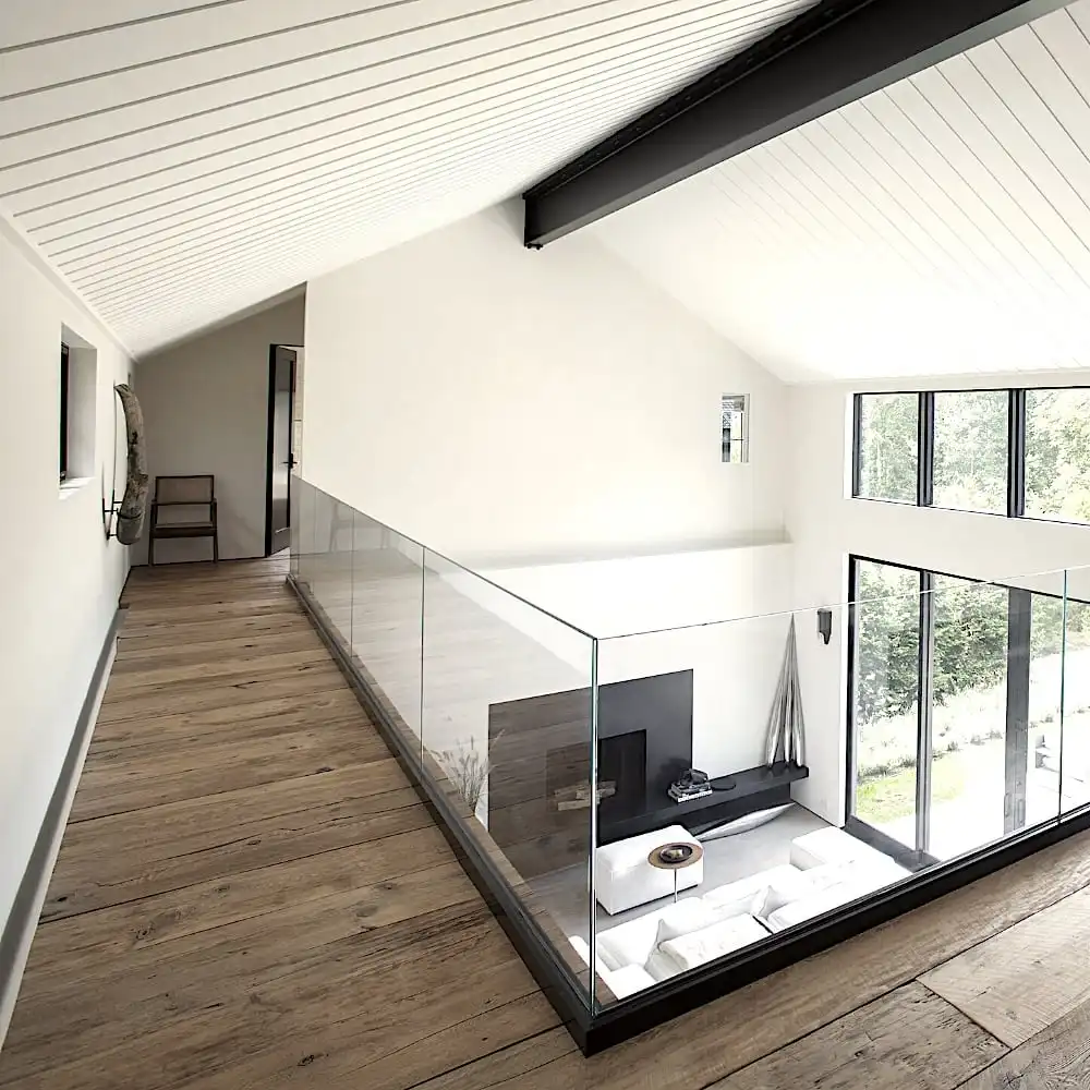  Un intérieur moderne comprend une passerelle au deuxième étage avec une balustrade en verre donnant sur un salon minimaliste. De grandes fenêtres apportent une lumière naturelle, mettant en valeur le parquet vieilli en sapin et la décoration contemporaine. 