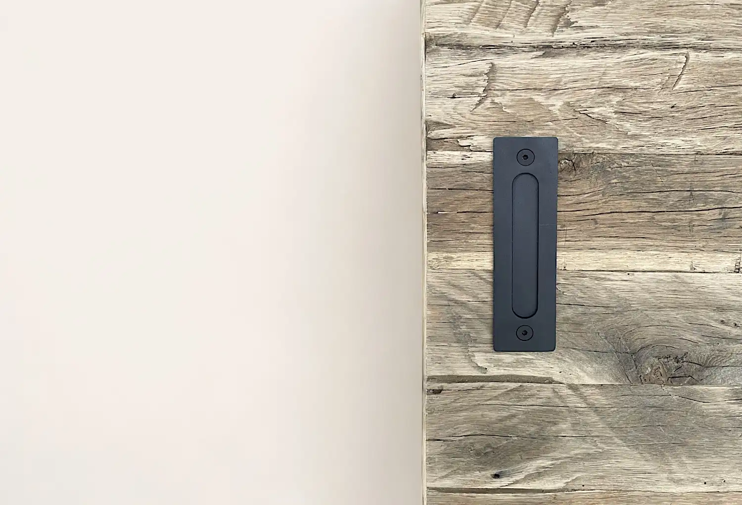 Une poignée de porte noire moderne est montée sur une surface en bois vieilli au grain texturé, adjacente à un mur blanc uni.