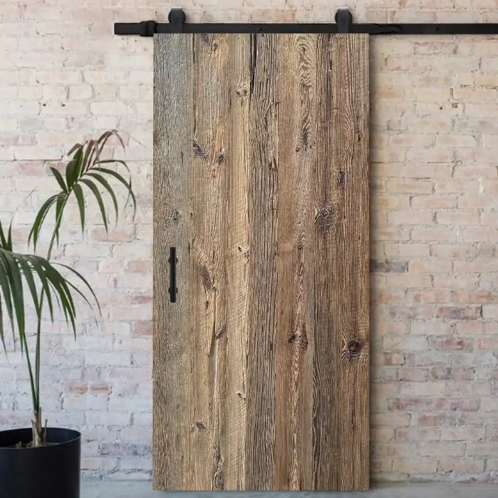  Une porte de grange coulissante avec une texture en bois rustique installée sur un rail noir, adossée à un mur de briques blanches dans une pièce avec une plante en pot à gauche. 