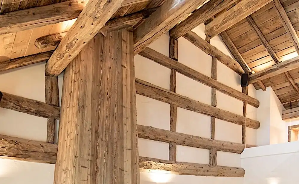 L'image montre l'intérieur d'un bâtiment avec des poutres en bois et des murs blancs, avec une construction rustique en bois et un plafond voûté avec du bois apparent, soulignant le charme des poutres anciennes de récupération.