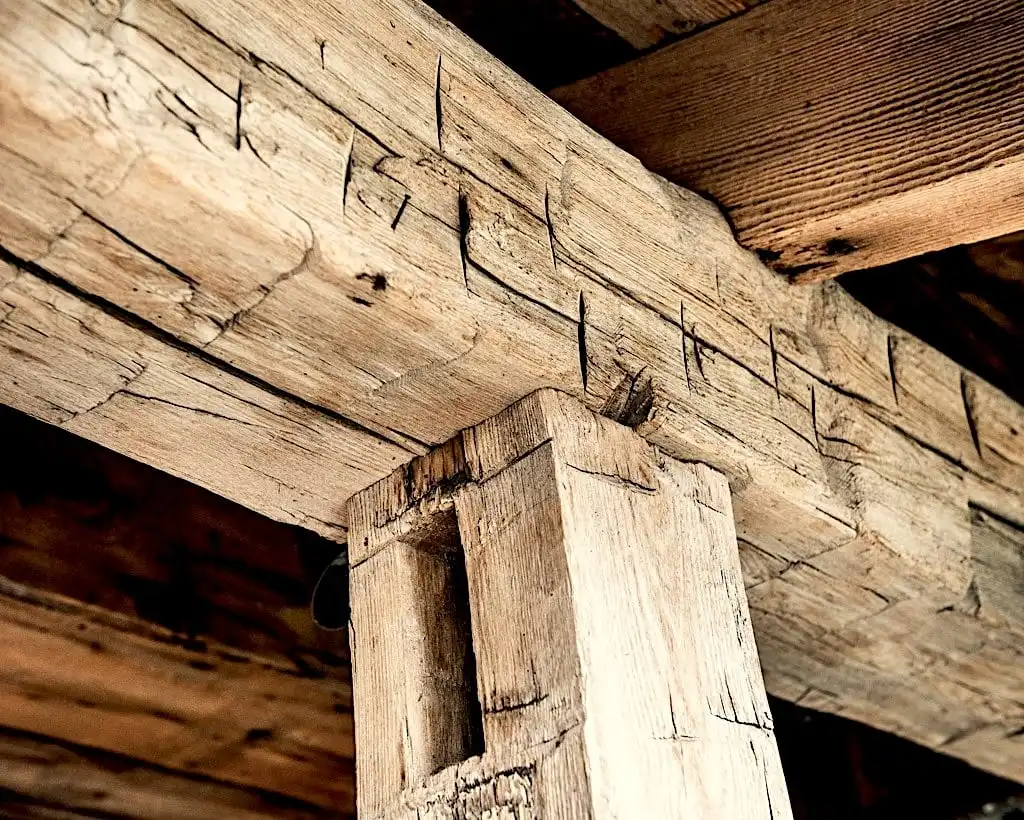 Gros plan d'une poutre et d'un poteau en bois rustique, présentant des fissures visibles et des textures naturelles, indiquant l'âge et l'usure des poutres anciennes de récupération.
