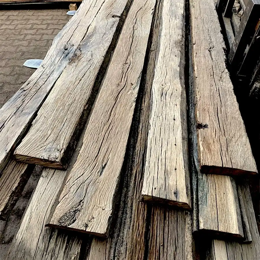  Plusieurs grandes poutres en bois patinées, comme des madriers anciens de chêne vieux, sont empilées les unes sur les autres à l'extérieur, avec des motifs de grains visibles et des textures rugueuses. 