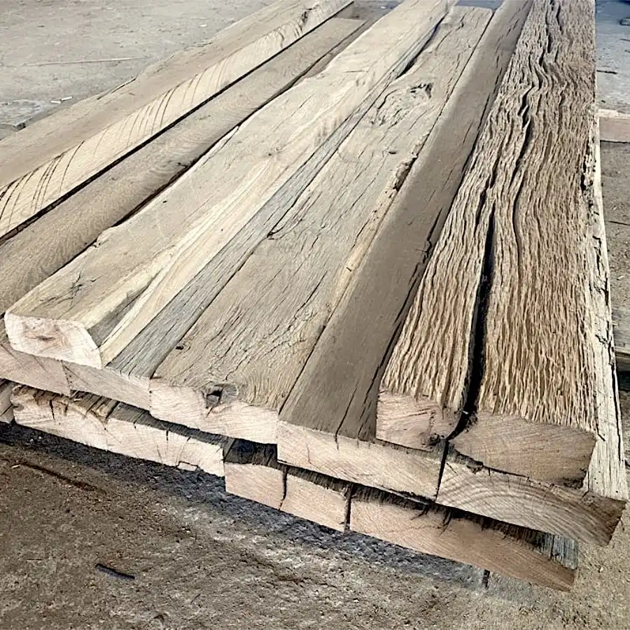  Empilement de planches de bois grossièrement taillées dans un décor d'atelier, présentant des textures et des longueurs variées, avec quelques madriers anciens ajoutant une touche de charme vintage. 