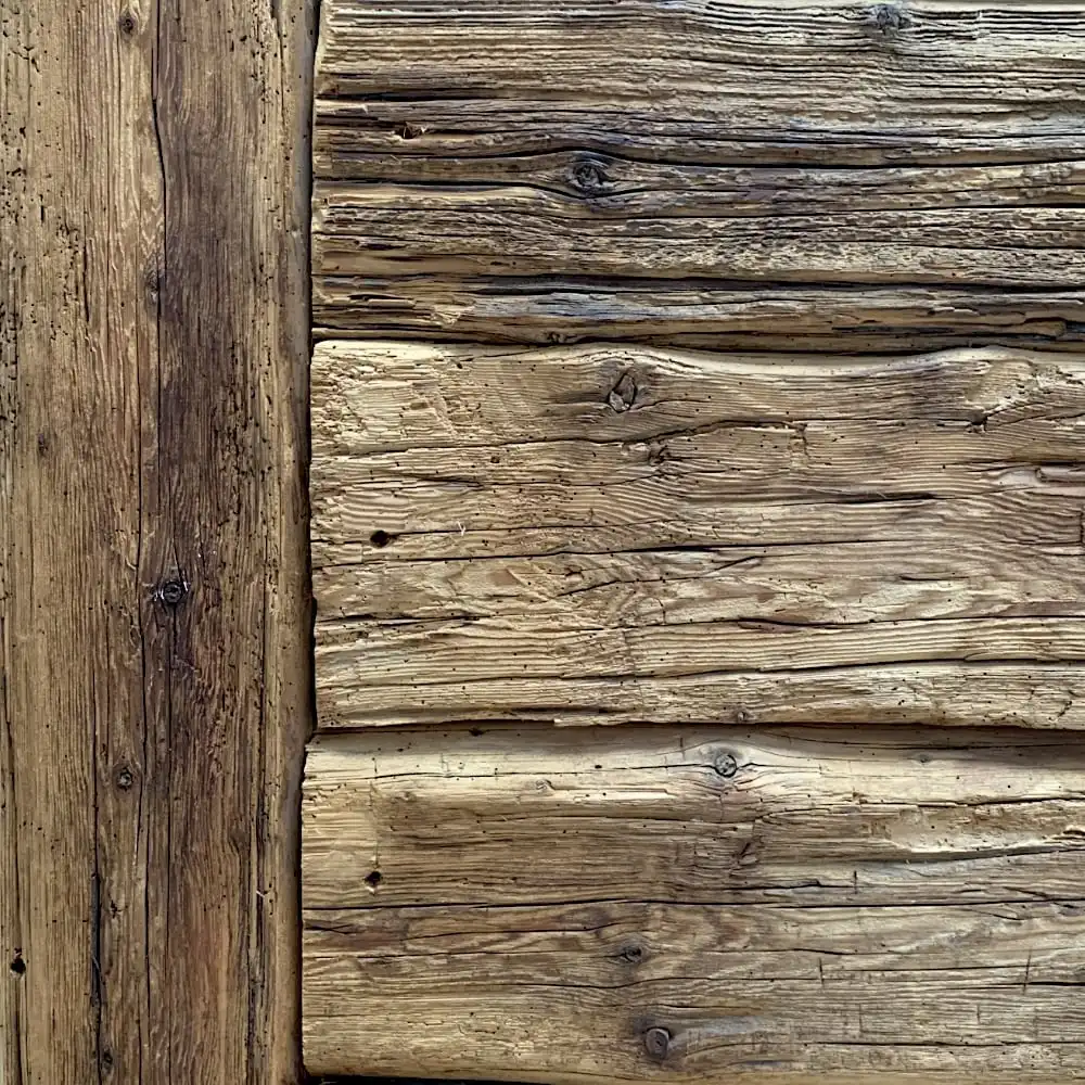  Gros plan d'un mur rustique en bois avec des rondins horizontaux montrant le grain et la texture naturels. Le bois semble patiné et vieilli, avec des nœuds visibles et des nuances de brun variées, un peu comme les madriers anciens. 