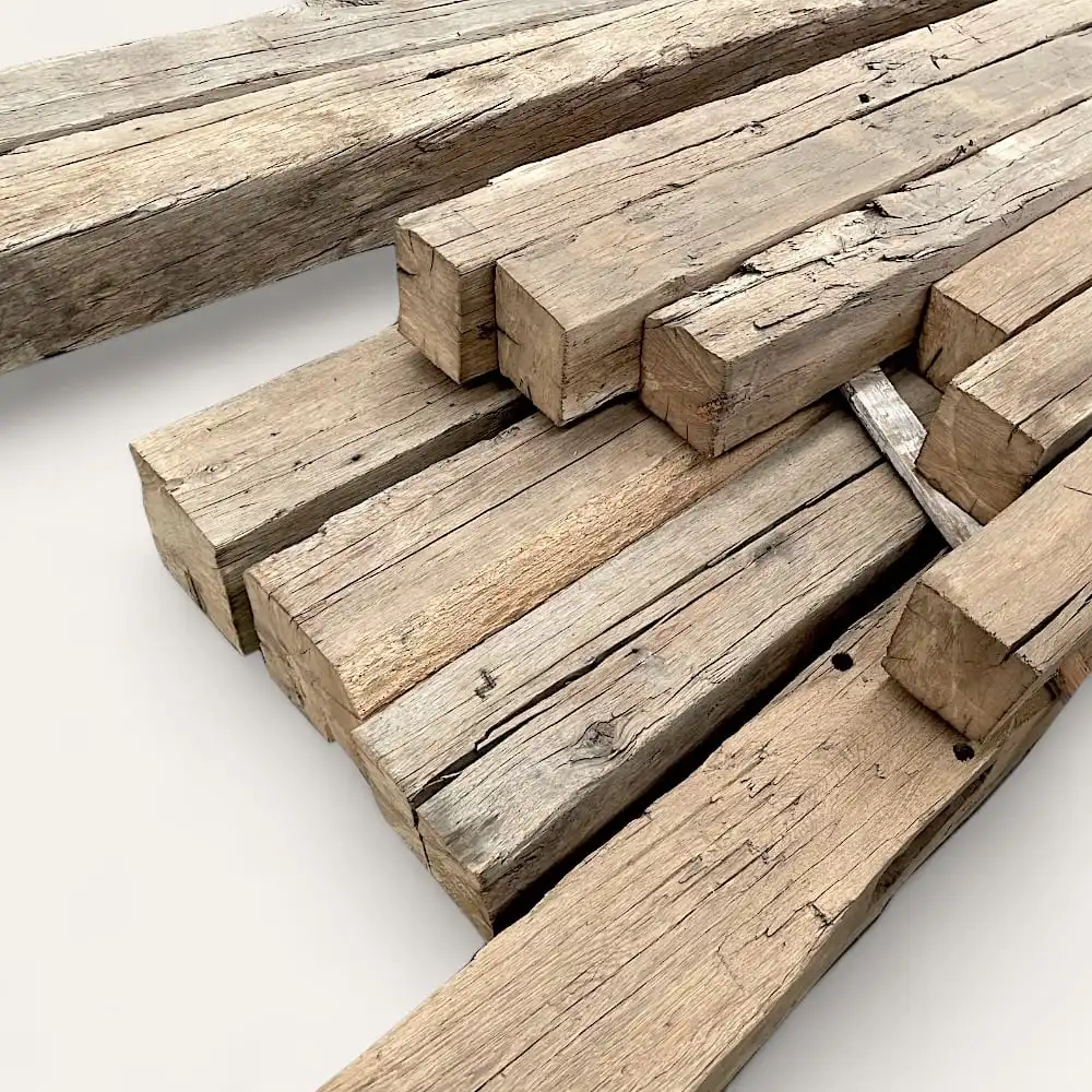  Un empilement de poutres en bois brut de différentes longueurs, rappelant les poutres anciennes de récupération, repose sur une surface plane. Le bois semble vieilli et altéré avec des fissures et des imperfections visibles. 