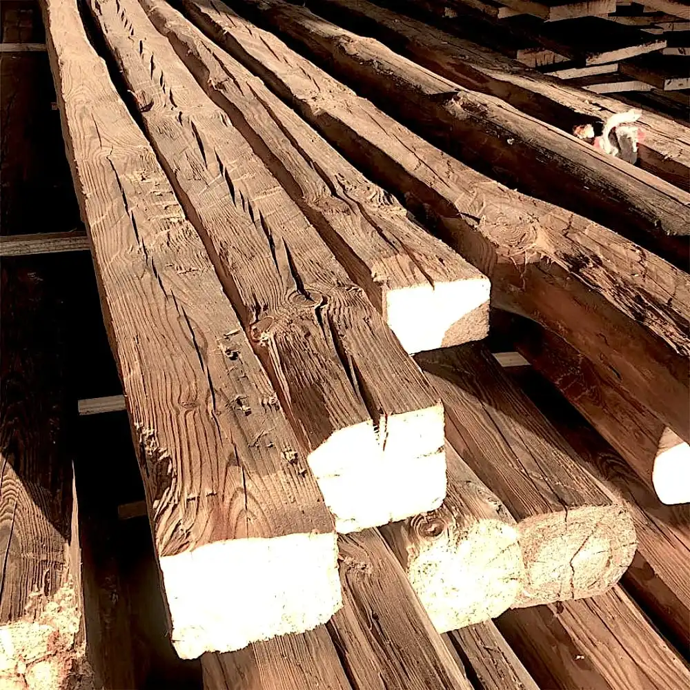  Plusieurs grandes poutres en bois grossièrement taillées empilées sur des supports, avec un chat se faufilant gracieusement entre les poutres anciennes de récupération en arrière-plan. 