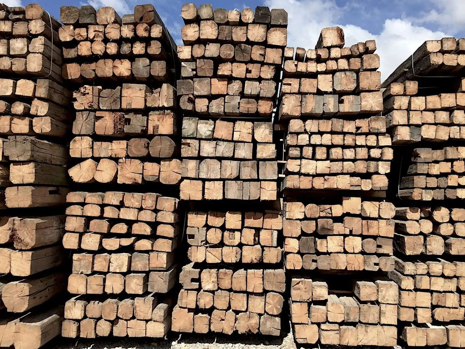 Des piles de morceaux de bois coupés de différentes tailles, y compris des poutres anciennes de récupération, soigneusement empilées à l'extérieur sous un ciel dégagé.