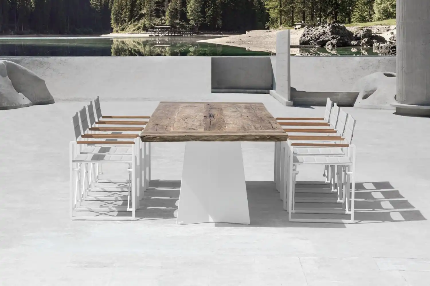 Ensemble de salle à manger extérieur moderne avec une table rustique en bois sur des pieds blancs trapézoïdaux et huit chaises blanches contemporaines, sur un patio en béton, avec vue sur un lac paisible et des arbres au loin.