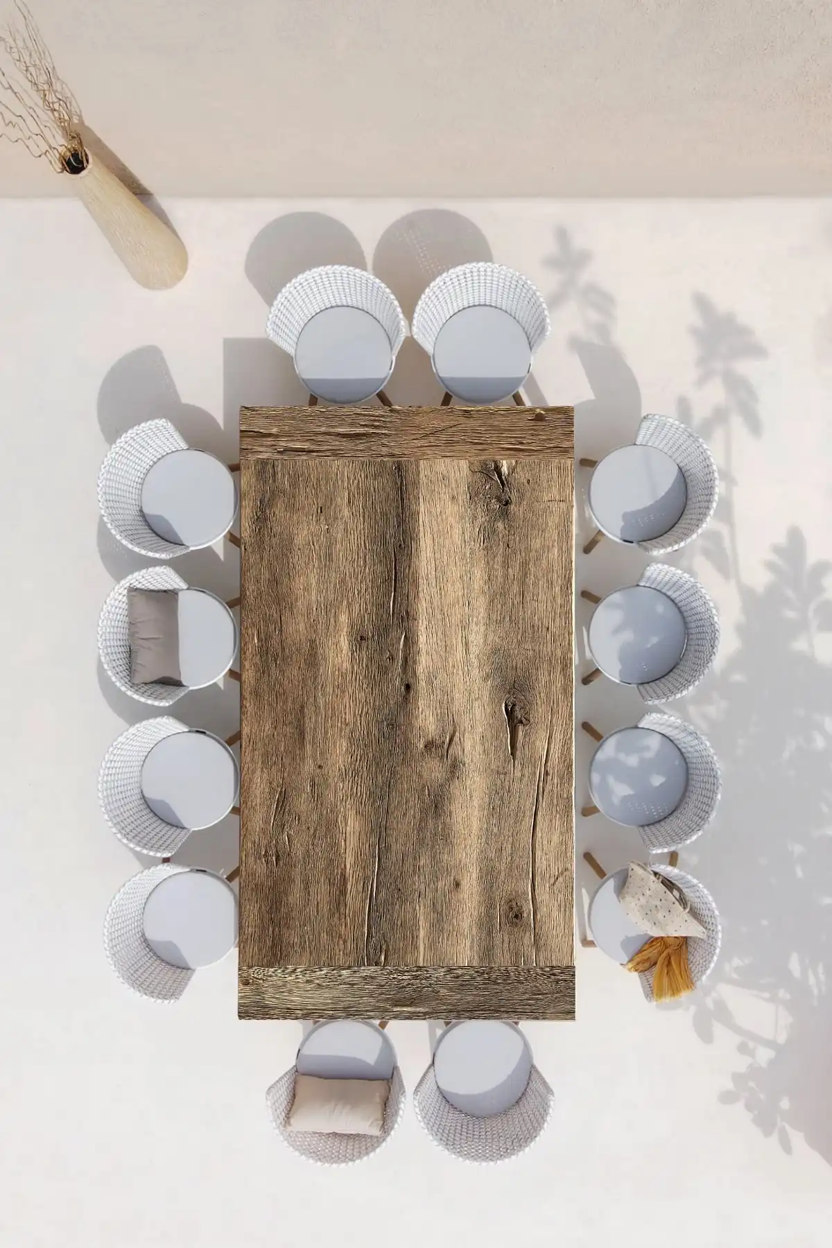 Vue aérienne d'une table rustique en bois brut disposée pour un repas avec des assiettes blanches texturées et des serviettes de table, le tout sur une nappe blanche épurée. Des ombres douces de feuillage se projettent sur la surface, évoquant une ambiance détendue en extérieur.