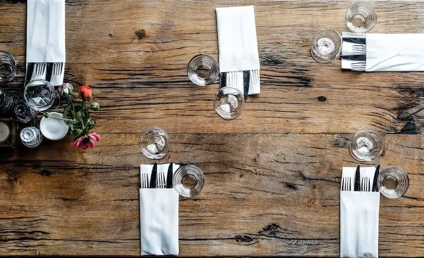 Vue de dessus d'une table en bois rustique dressée pour quatre, avec des serviettes, des couverts, des verres et un petit bouquet central.