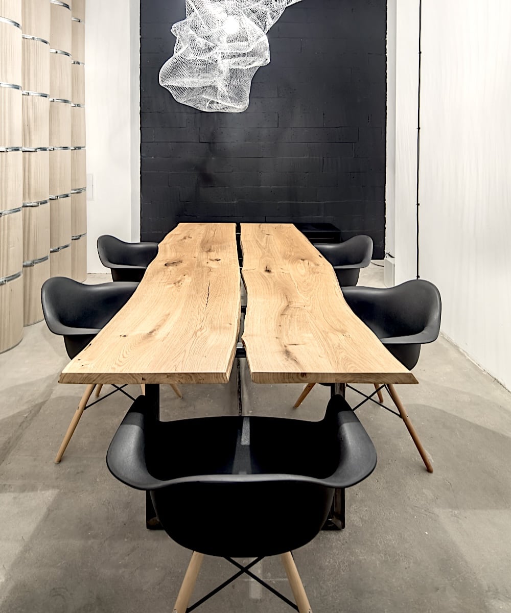 Une table de style rustique avec un plateau en bois brut à deux planches séparées par un espace, reposant sur des pieds en métal noir. Autour de la table se trouvent des chaises design noires avec des pieds en bois et métal.