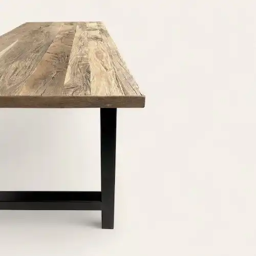  Coin d'une table rustique avec plateau en bois vieilli et pied en métal noir mat, contrastant avec le fond uni et lumineux qui met en valeur son design épuré et moderne. 