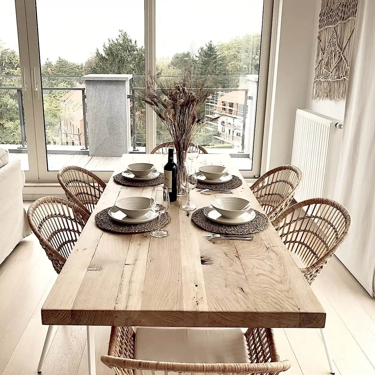  Salle à manger lumineuse avec grande table en bois ancien dressée pour un repas, entourée de chaises en rotin, décoration naturelle et vue extérieure. 