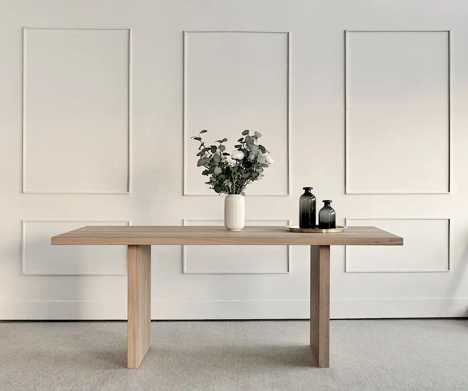  Console minimaliste en bois clair avec décoration épurée, vase blanc avec eucalyptus et bouteilles noires, sur fond de mur blanc avec cadres décoratifs. 