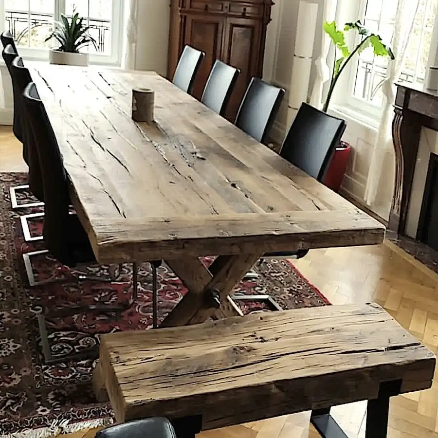  Grande table de style fermette en bois ancien robuste, présentée dans un intérieur classique avec un tapis persan, des chaises modernes et des touches de verdure, reflétant une ambiance chaleureuse et accueillante. 