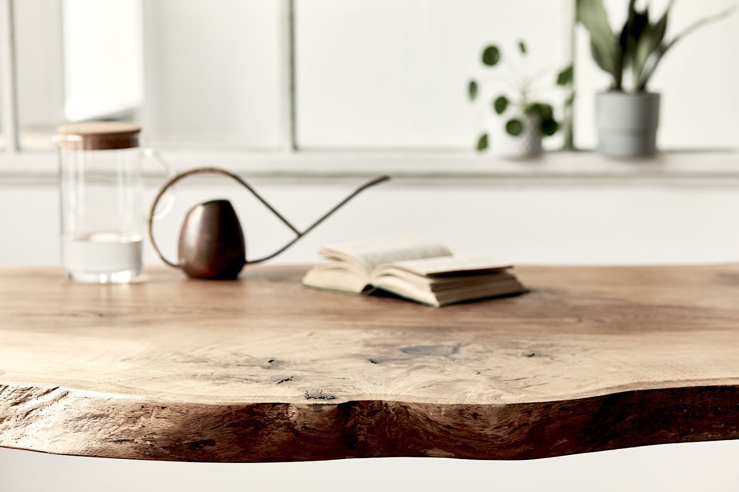 Une table live edge en bois avec un bord irregulier contient une carafe en verre, un arrosoir et un livre ouvert. Des plantes en pot sont visibles sur le rebord de la fenêtre en arrière-plan.