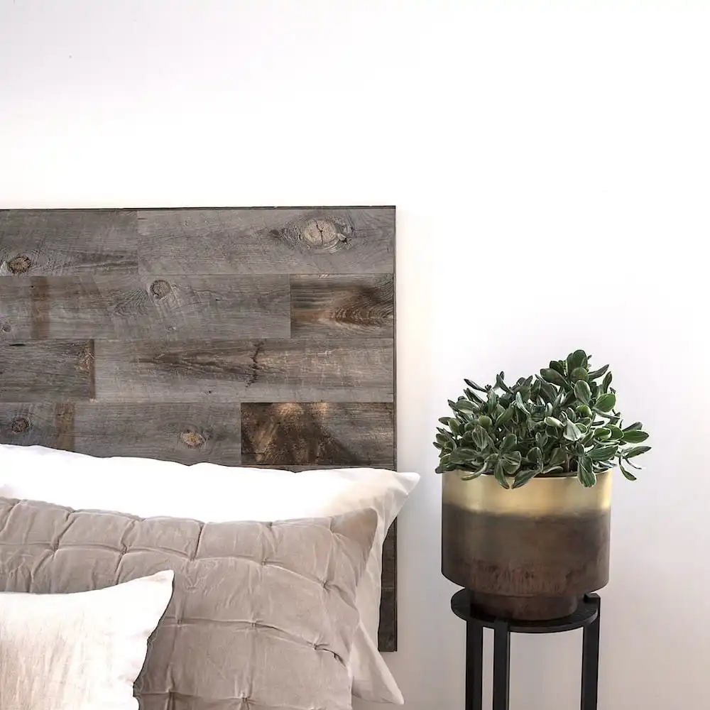  Un coin chambre moderne avec une tête de lit en bois ancien, des oreillers blancs, une couette grise et une plante en pot sur un support noir. 